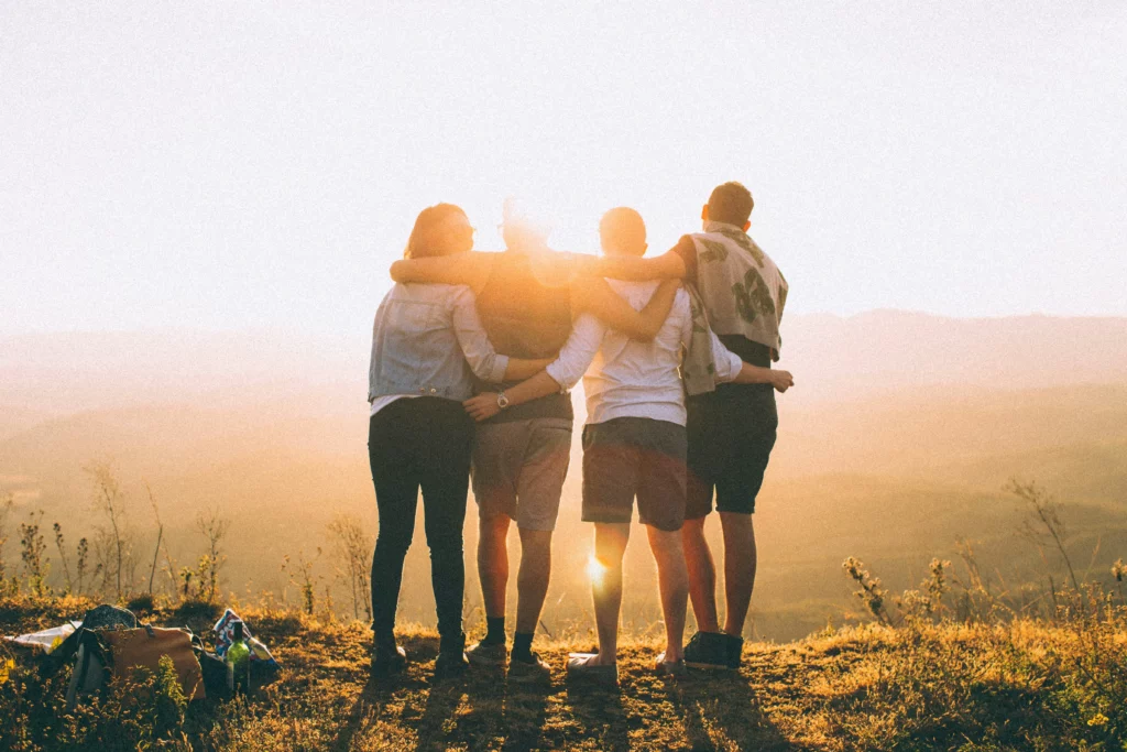 Cette image illustre la page "Qui suis-je ?". On y voit un groupe d'amis debout, côte à côte, se tenant amicalement par la taille ou les épaules face à  un coucher de soleil.